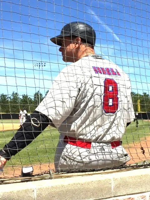 A baseball player in a white and gray uniform with the number 8 stands behind a fence, viewed from the back, looking out onto a sunny baseball field.